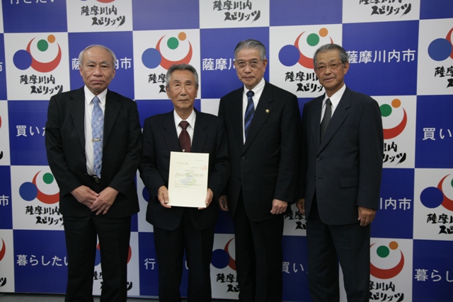 4人のスーツ姿の男性4人が並び、向かって左から2番目の人物が書類を持っている写真