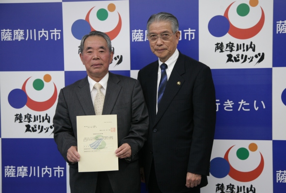 二人の男性が並び、左側に立っている人物が書類を持っている写真