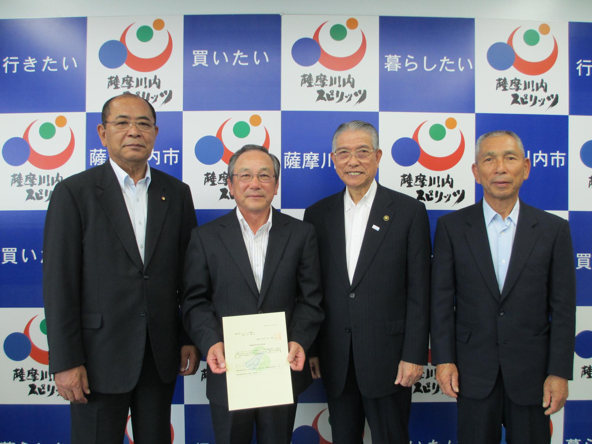 4人のスーツ姿の中高年の男性が並び、左から2番目の人物が書類を手に持っている写真