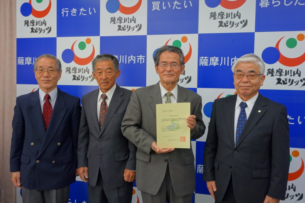 4人の中高年のスーツ姿の男性が並び、右側から2番目の人物が書類を持っている写真