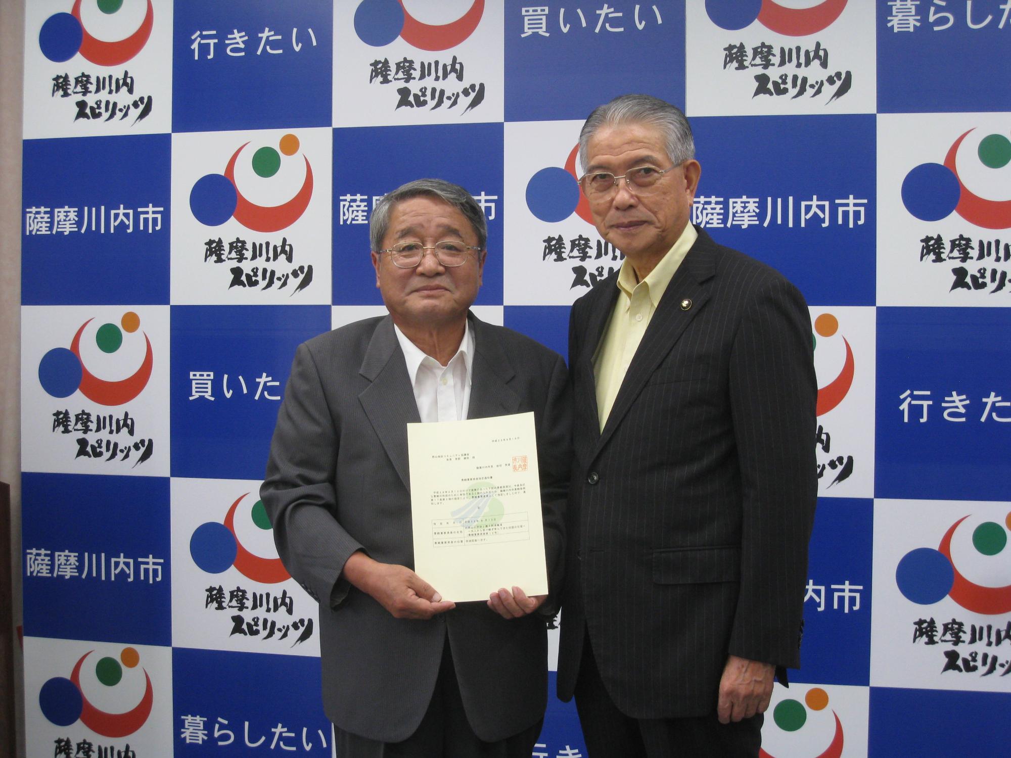 中高年の男性2人がスーツを着て並び、左の人物が書類を持っている写真