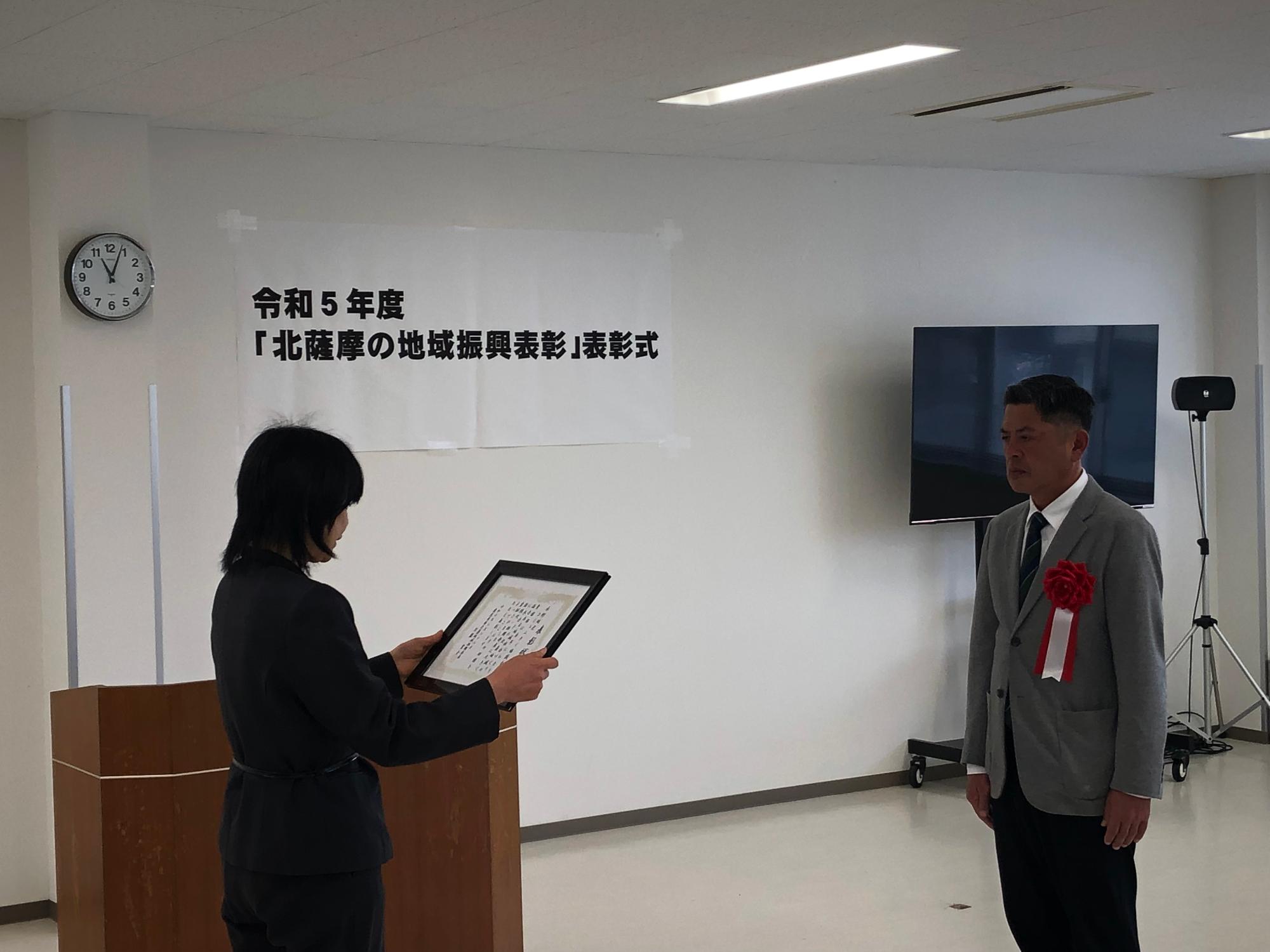 令和5年度「北薩摩の地域復興表彰」表彰式で、胸に赤い造花を付けたスーツ姿の男性が北薩地域振興局長から表彰状を受け取る写真