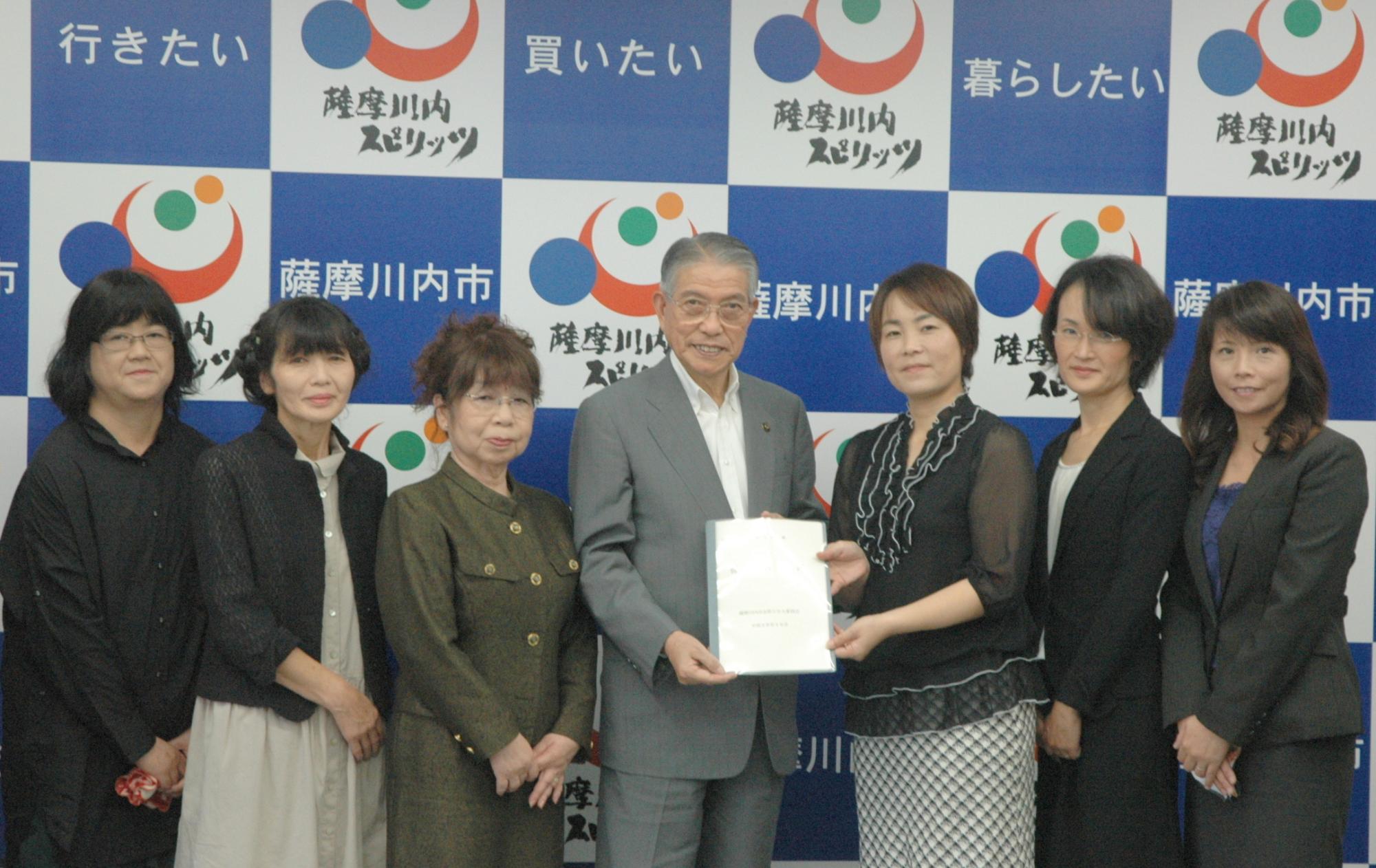 薩摩川内市と書かれたバックボードを背に、スーツを着たロマンスグレーの髪持つ一人の男性とその男性を囲むように左方と右方それぞれに三人ずつご婦人が立ち、中心にいる男性と女性が一緒に提言書を掲げている写真