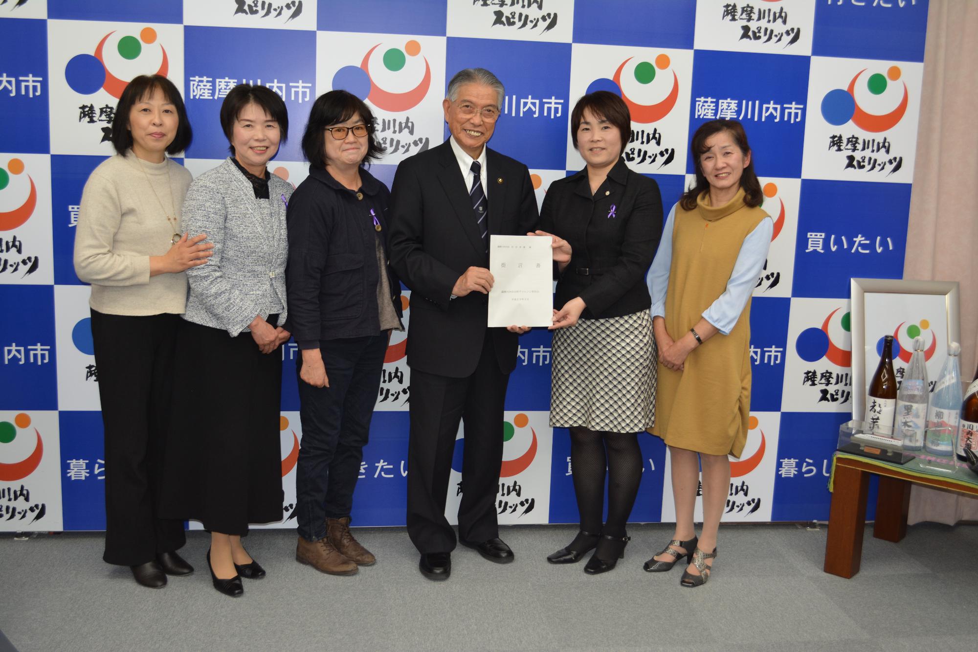 薩摩川内市女性チャレンジ委員会の5名と市長が、薩摩川内市と書かれたパネルの前で横一列に並んでいる写真。薩摩川内市女性チャレンジ委員会が取りまとめた事業構想を、右から2番目の女性が、左隣の市長に手渡している様子が撮影されている。