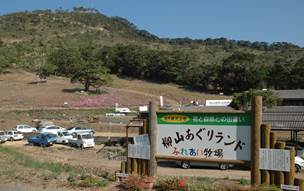 手前に柳山あぐりランドふれあい牧場と書かれた看板が立っており、その少し奥に10台近くの車が止まっている小さな駐車場があり、さらに奥には見晴らしのよさそうな丘が映っている写真