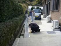 コミュニティセンターと生垣の間にある通路で、三人の業者が通路に軽トラックを置きグレーチングを設置している写真