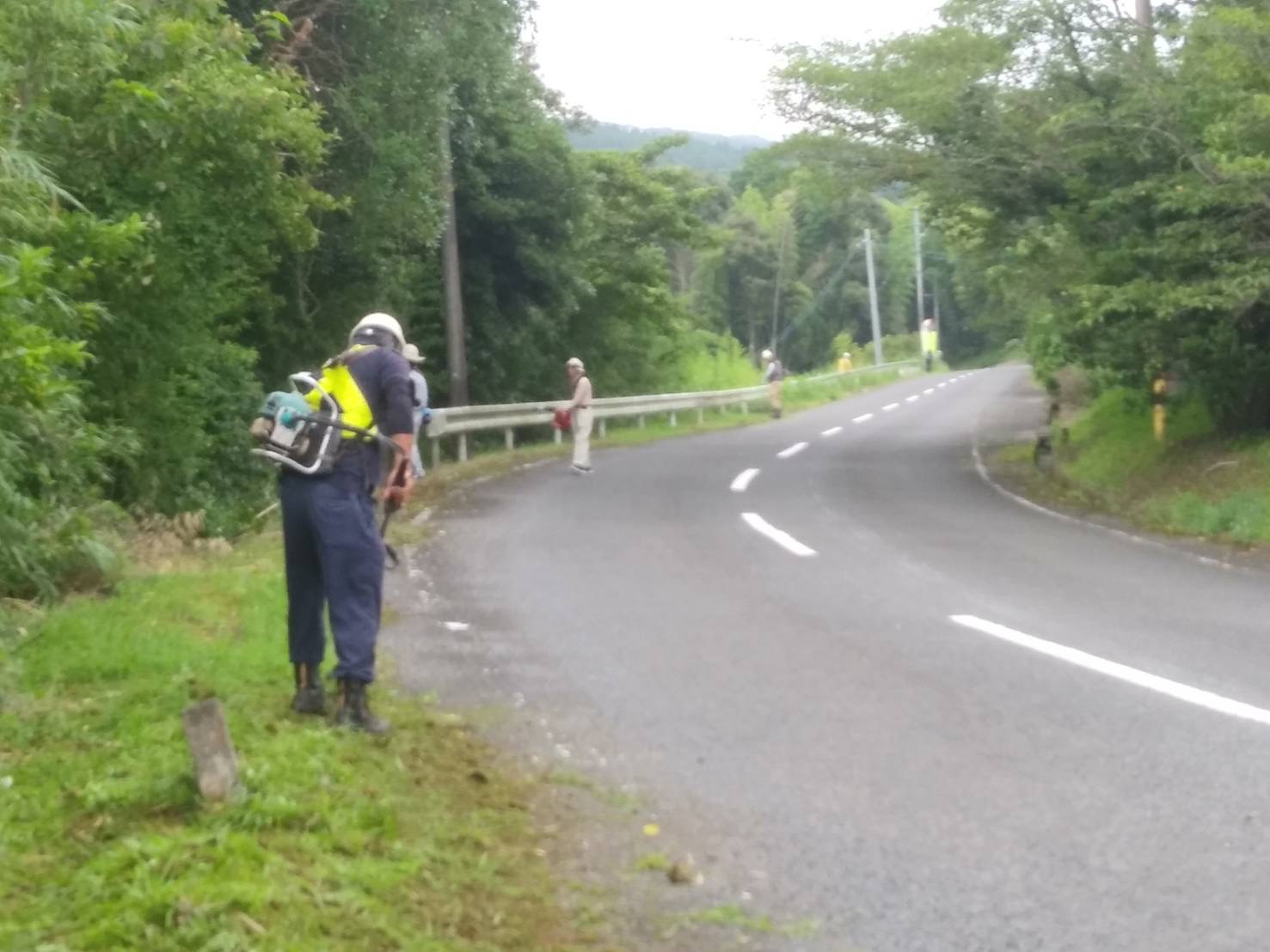 八重自治会(八重地区)の道路脇で、4名の男性作業員が草払い機で約10メートル間隔で草払いをしている写真。