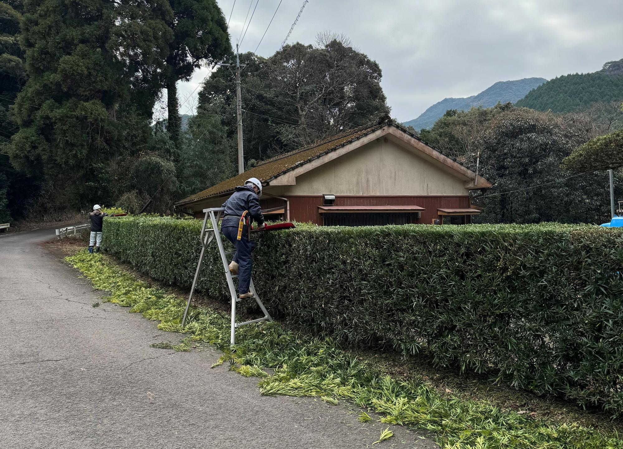 矢立自治会(黒木地区)の公民館で、2名の男性作業員がチェンソーで樹木剪定をしている写真。