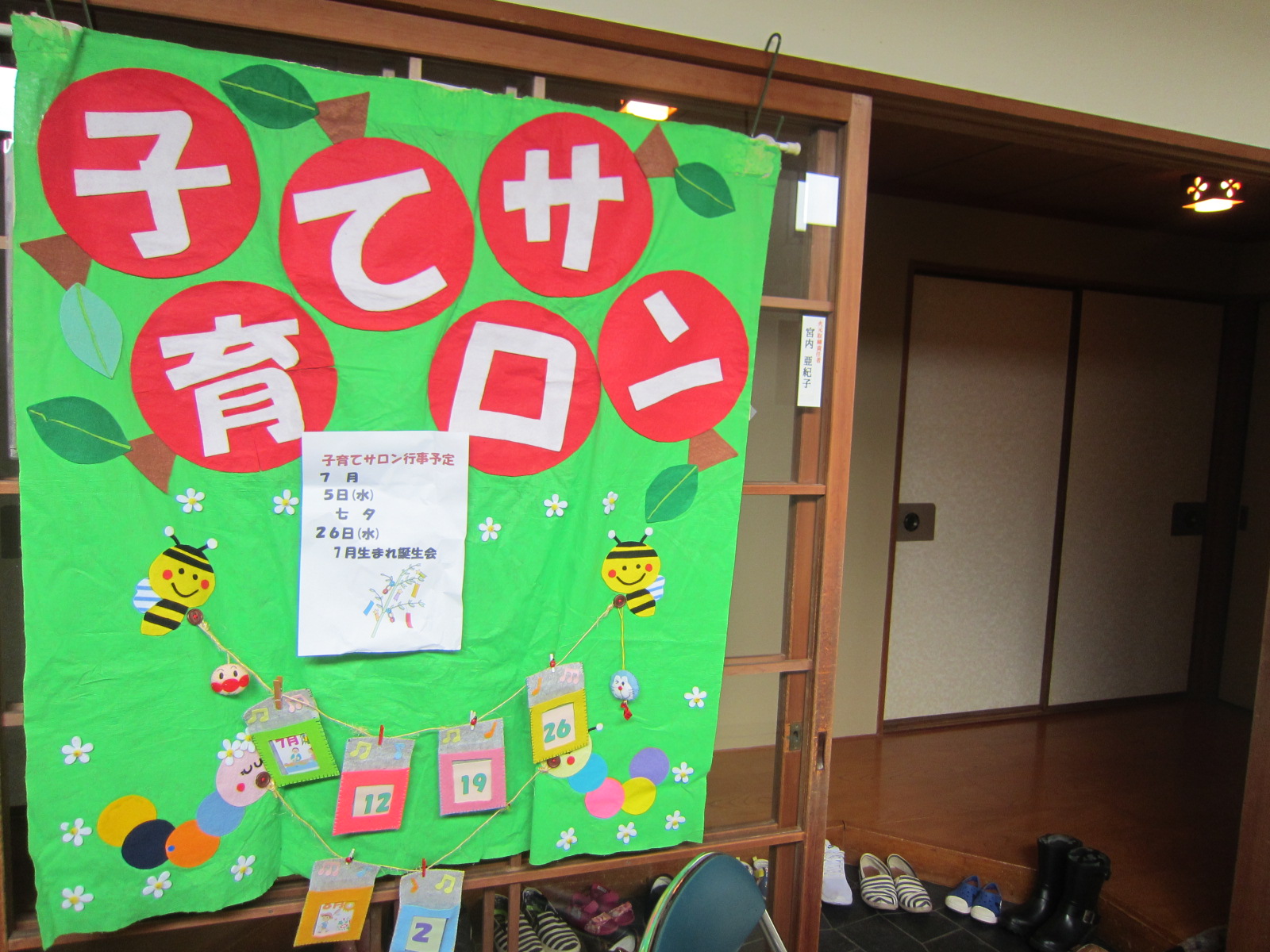 「子育てサロン」と書かれている文字の下に、蜂が日付の書かれた紙をぶら下げているイラストで作られた横断幕が壁に垂れ下がってる写真
