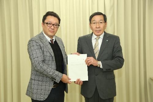 グレーのチェックのブレザー姿の男性とグレーのスーツ姿の男性が一つの書類を一緒に持ち前を向いている写真