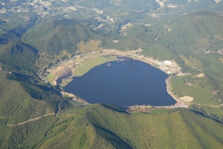 緑に覆われた山に囲まれた青黒い色の大きな池を上空から撮った写真