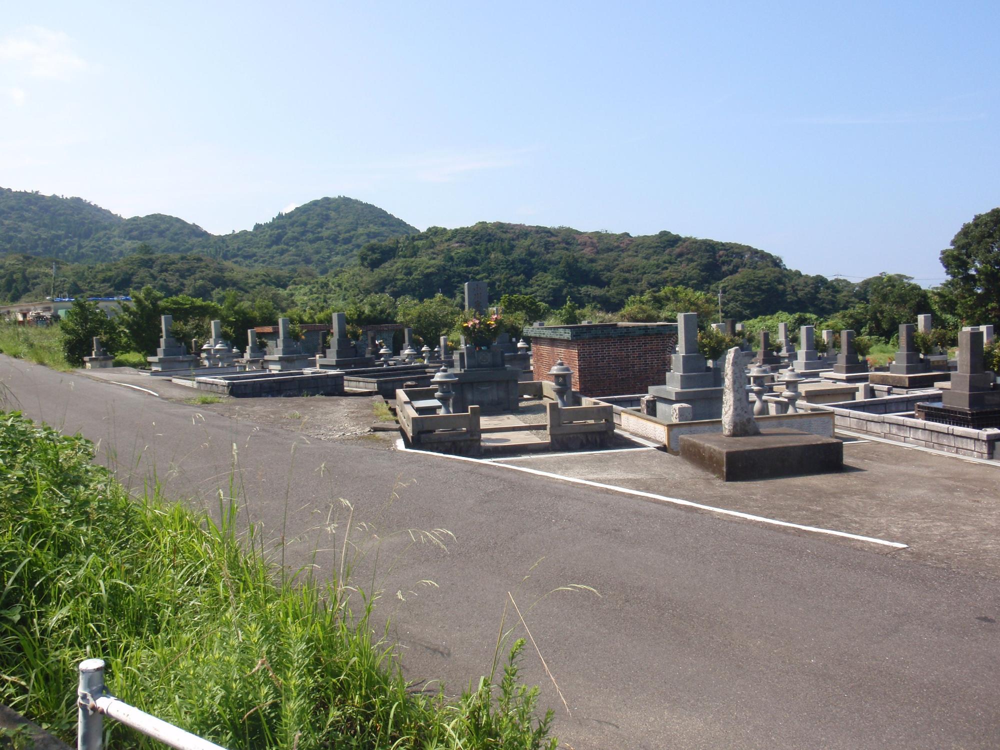 青空の下、道路に面した数基のお墓の後ろに、緑の山々が写っている写真