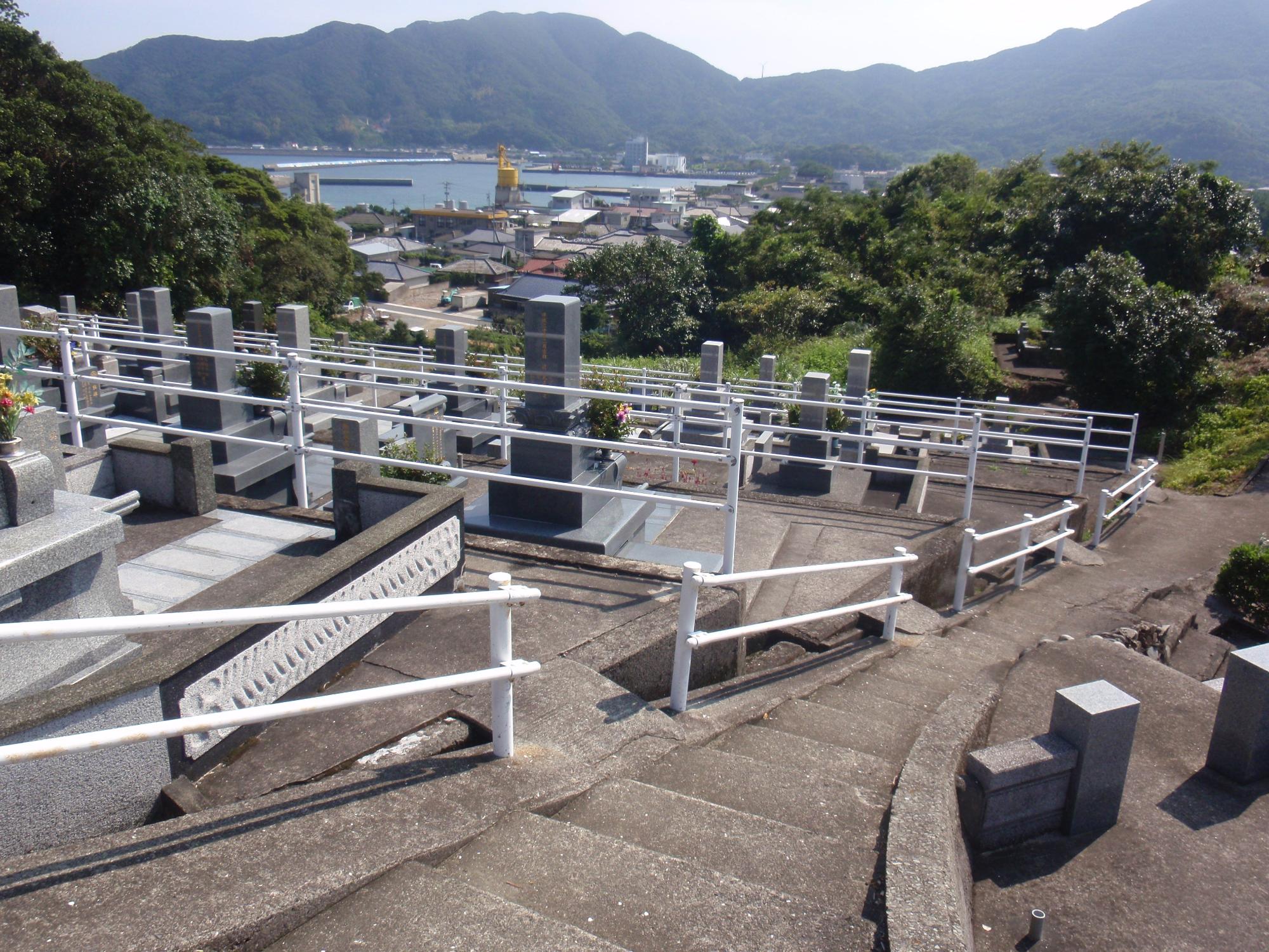 青空の下、細い階段の脇に段々になって建つ数基のお墓から港を見下ろし、対岸に山が広がっている景色の写真