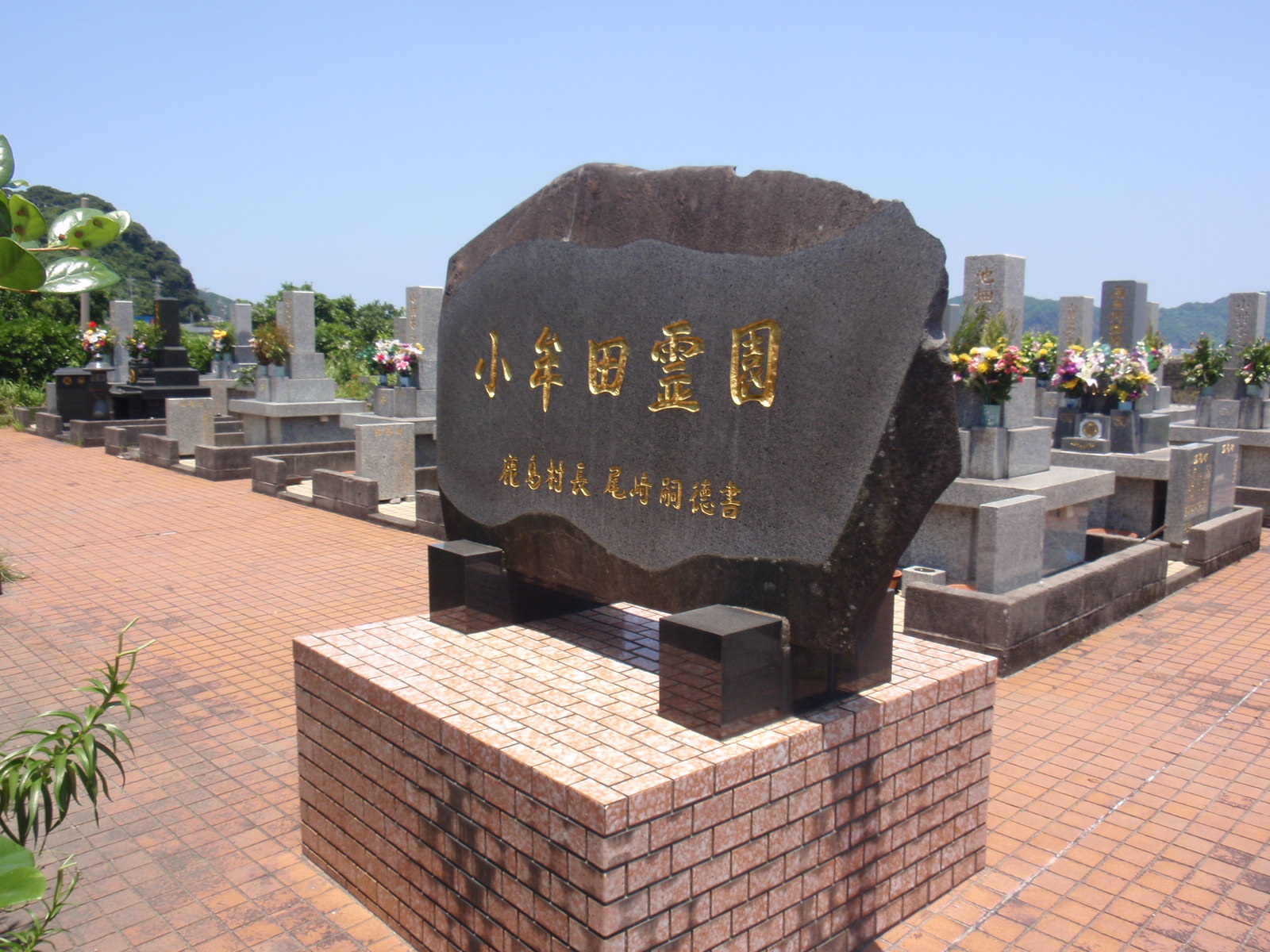 青空の下で整然と並ぶ花をお供えされたお墓を背景に、金色の文字で小牟田霊園と書かれた大きな黒い石が建てられている写真