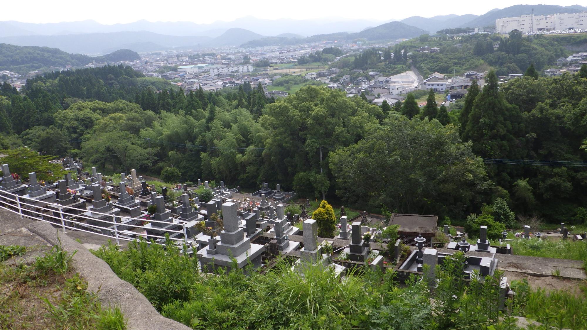 斜面に並ぶお墓を見下ろし、墓地の先に町が広がり、町の先に山々が霞がかって見えている写真