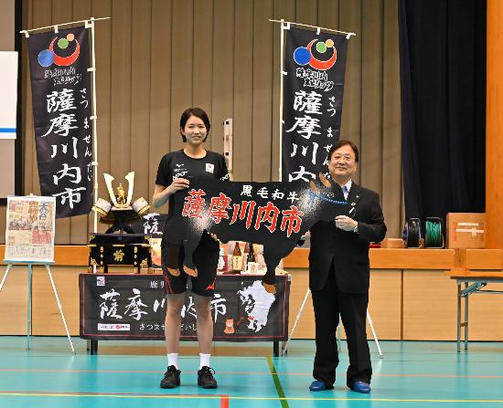 キャプテンの古賀紗理那選手と並んで特産品を手に写真に写る田中市長