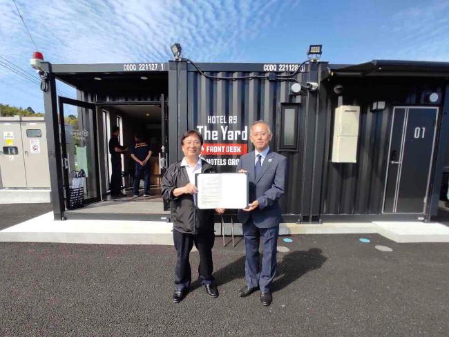 協定書を持ちコンテナを背に写真に写る田中市長