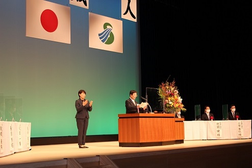 日の丸と薩摩川内市の旗が掲げられている講堂で祝辞を述べている市長とそれを手話にしている女性と関係者たちの写真
