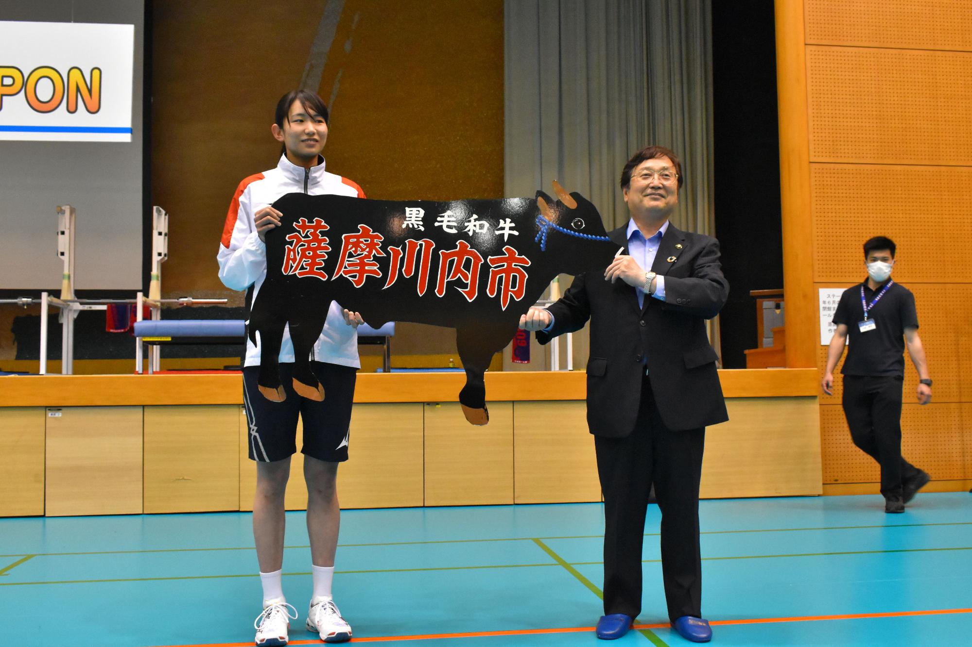 黒毛和牛、薩摩川内市と書かれた黒い牛の形の目録を掲げている、市長と女子バレーボール日本代表チームのメンバーの写真