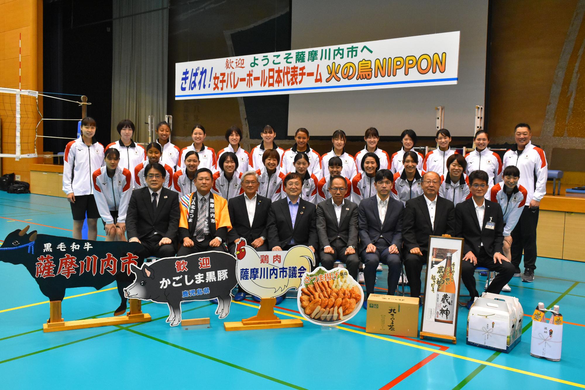 吊り看板の下、8種類の目録を前に市長と関係者と日本代表チームが収まっている記念写真