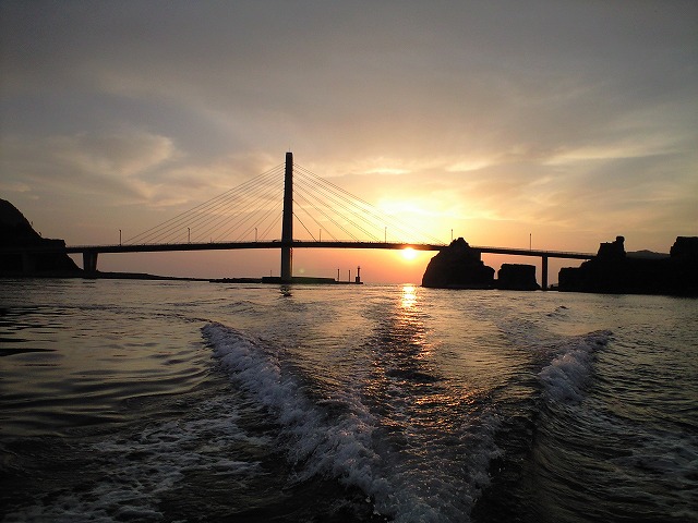 海にかかる橋と白波の立つ海面ににオレンジ色の空から太陽の光が差している写真