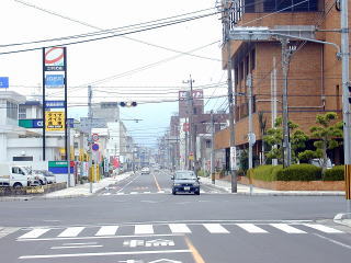 左側にガソリンスタンドがあり右側に茶色い建物が建っている間の道路を車が走っている写真