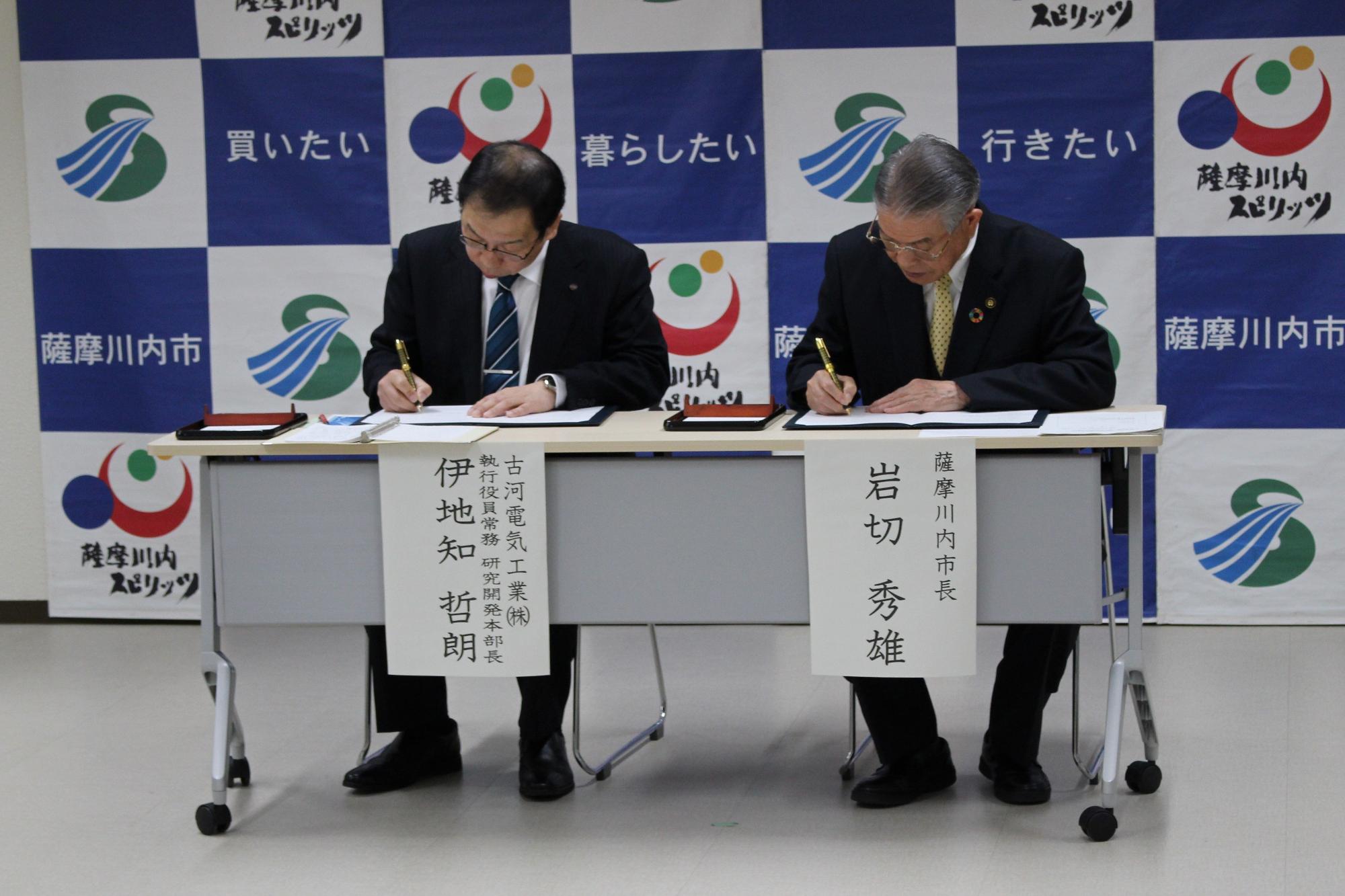 スーツ姿の男性二人が同じ机に並んで書類にサインしている写真