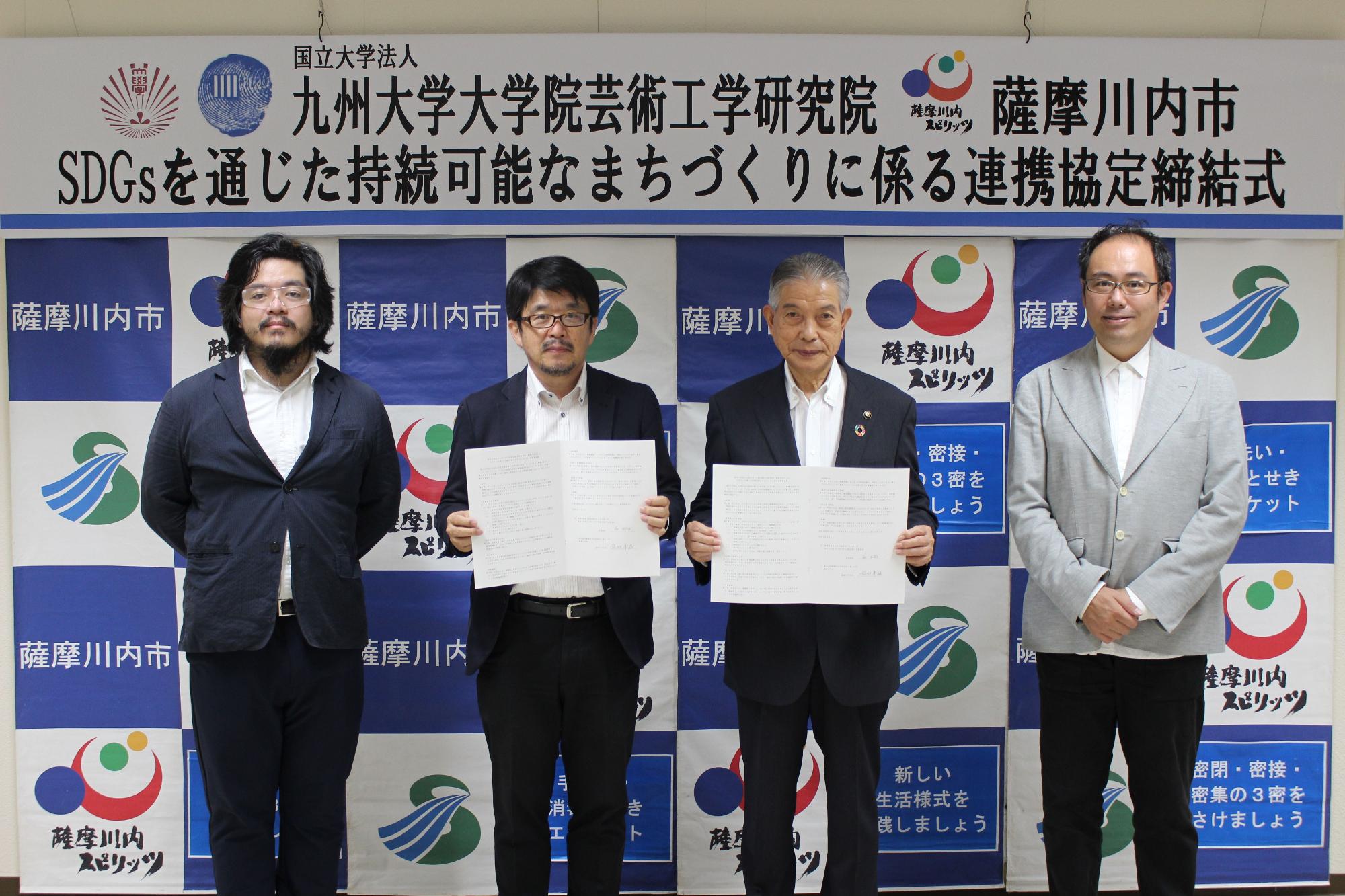 「SDGsを通じた持続可能なまちづくりに係る連携協定締結式」と書かれた横断幕の下で、書類を掲げるスーツ姿の4人の男性たちの写真