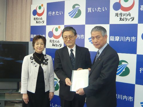 スーツ姿の男性2人が書面を一緒に持ち、その隣に女性が並んでいる記念写真