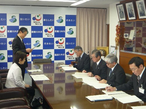 男性数名と女性がそれぞれ長テーブルに座って置かれた書面を確認する中、奥の男性が立って書面を読み上げている写真