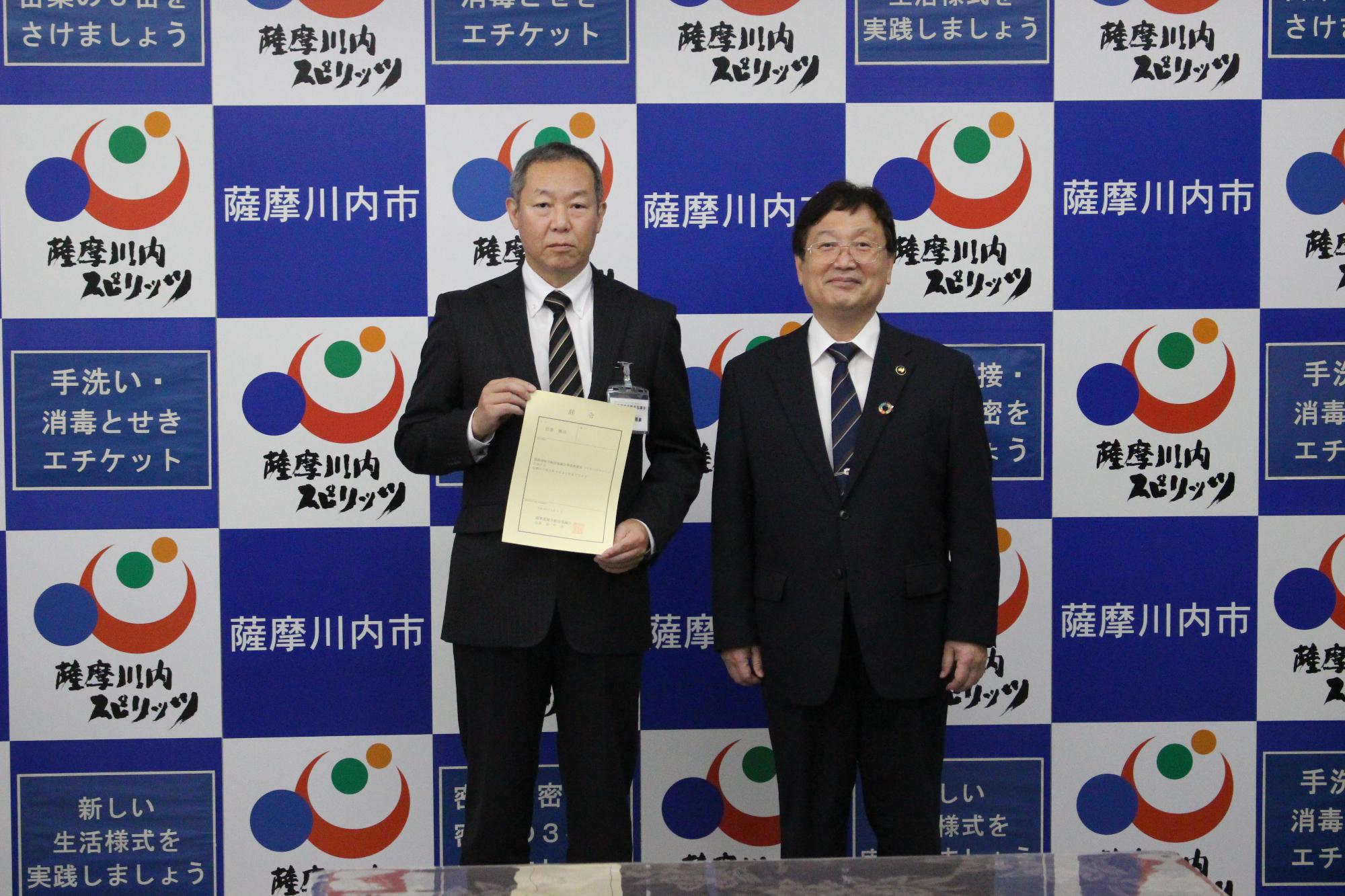 薩摩川内市等と書いてある壁紙の前で、二人のスーツ姿の男性が立っている写真