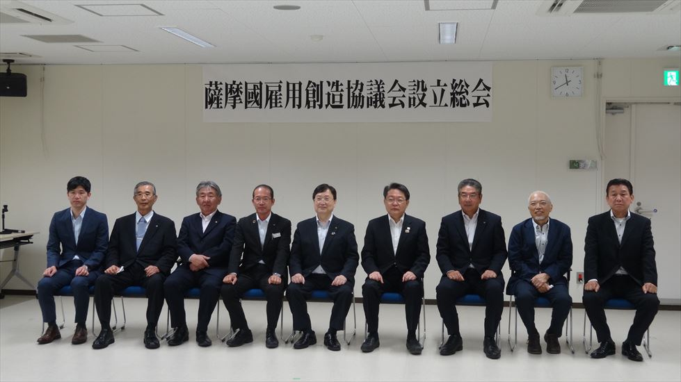 薩摩國雇用創造協議会設立総会と書かれた看板の前に横一列で座るスーツ姿の男性たちの写真