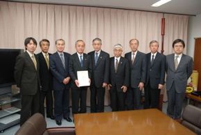 9人のスーツを着た男性が並び、向かって左から4番目の人物が書類を手に持っている写真