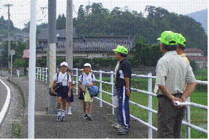 隣に田んぼ見える歩道を歩いている男子小学生と、それを見守っている黄色いつば付き帽子をかぶった3人の年配男性の写真