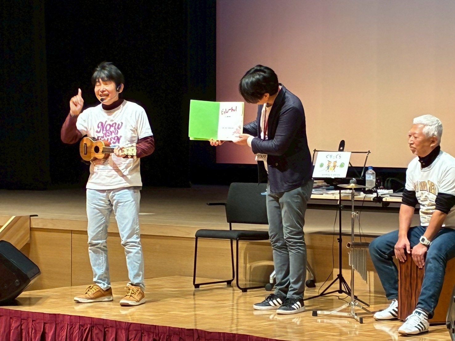 絵本ライブで3人の男性が右から座っている人、絵本を持っている人、楽器を片手に説明している人が映っている写真