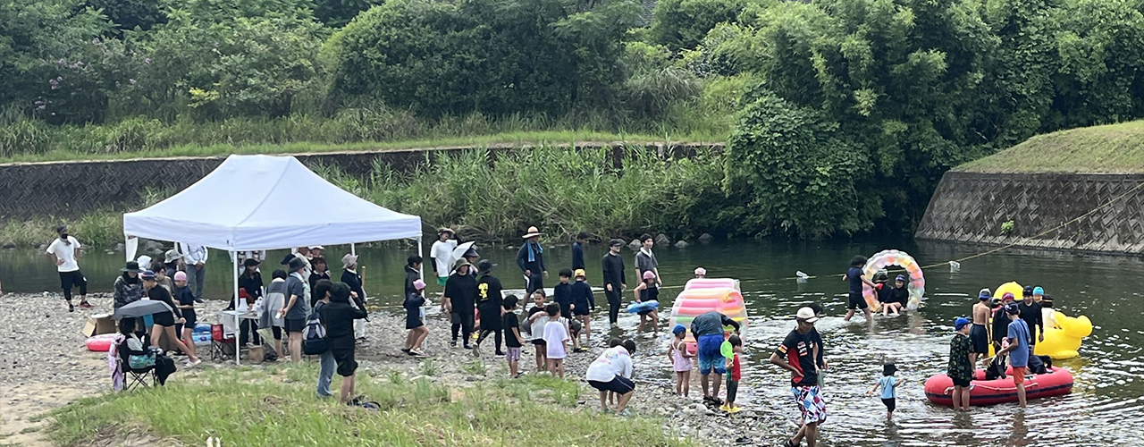 川辺で十数名がテントを張ったり浮き輪を用いて遊んでいる様子