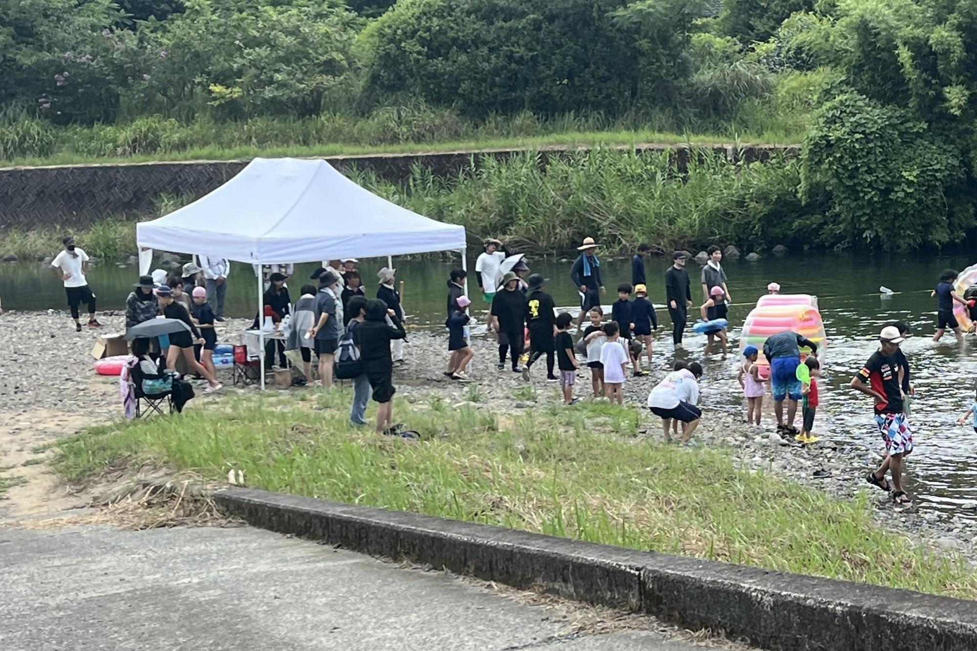川辺で十数名がテントを張ったり浮き輪を用いて遊んでいる様子