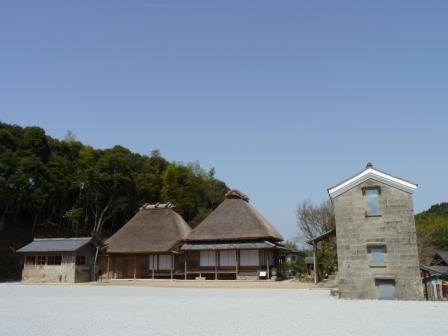 茅葺屋根の建物と、その左右に建物があり、奥に木々が見える写真