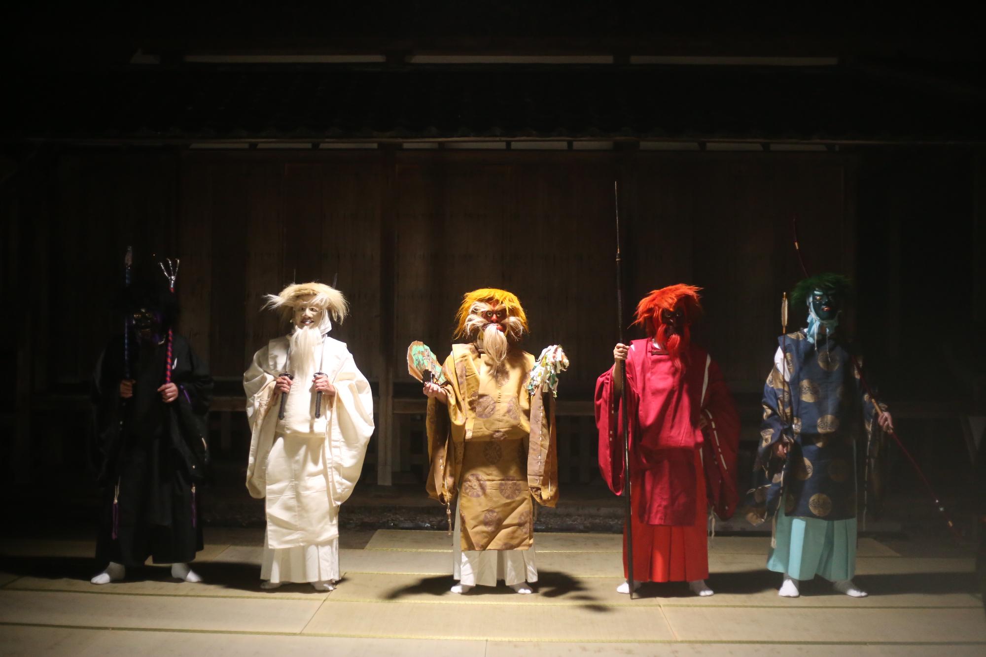 薄暗い室内に立つ、和装を着て面を被った演者たちの写真