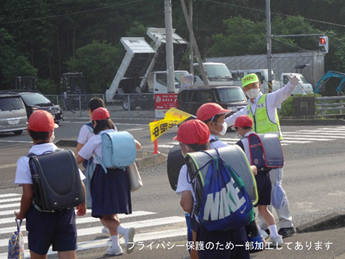 赤い帽子を被り横断歩道を渡る児童と黄緑のビブスを着用し横断旗を振る少年愛護員の写真（プライバシー保護のため一部加工してあります）