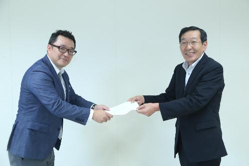 白い壁を背景にスーツを着た二人の男性が向かい合い一つの書類を互いに持ち合っている様子の写真