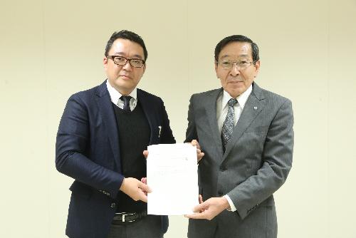 壁をバックにスーツを着た二人の男性が正面を向き一つの書類を一緒に持っている写真
