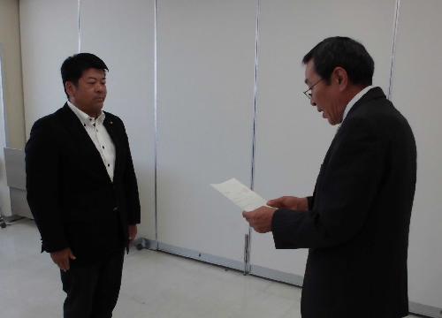 眼鏡を掛けたスーツ姿の男性が正面に立つスーツ姿の男性に向かい書面を読み上げている様子の写真