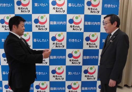 薩摩川内スピリッツと書かれたパネルの前でスーツ姿の2人の男性が向かい合い書面の受け渡しをしている様子の写真
