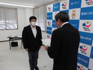 長机と椅子が置かれた室内にてパネルを背景にスーツを着た二人の男性が向き合って書類の受け渡しを行っている様子の写真