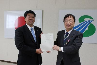 スーツを着た二人の男性が日本国旗と薩摩川内市市章を背景に並んで立ち正面を向きながら一つの書類を互いに持ち合っている写真